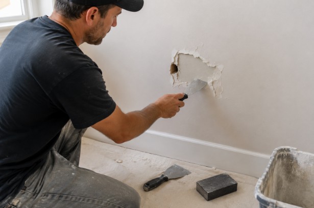 A workman scraping loose drywall from a damaged hole in a wall with a putty knife